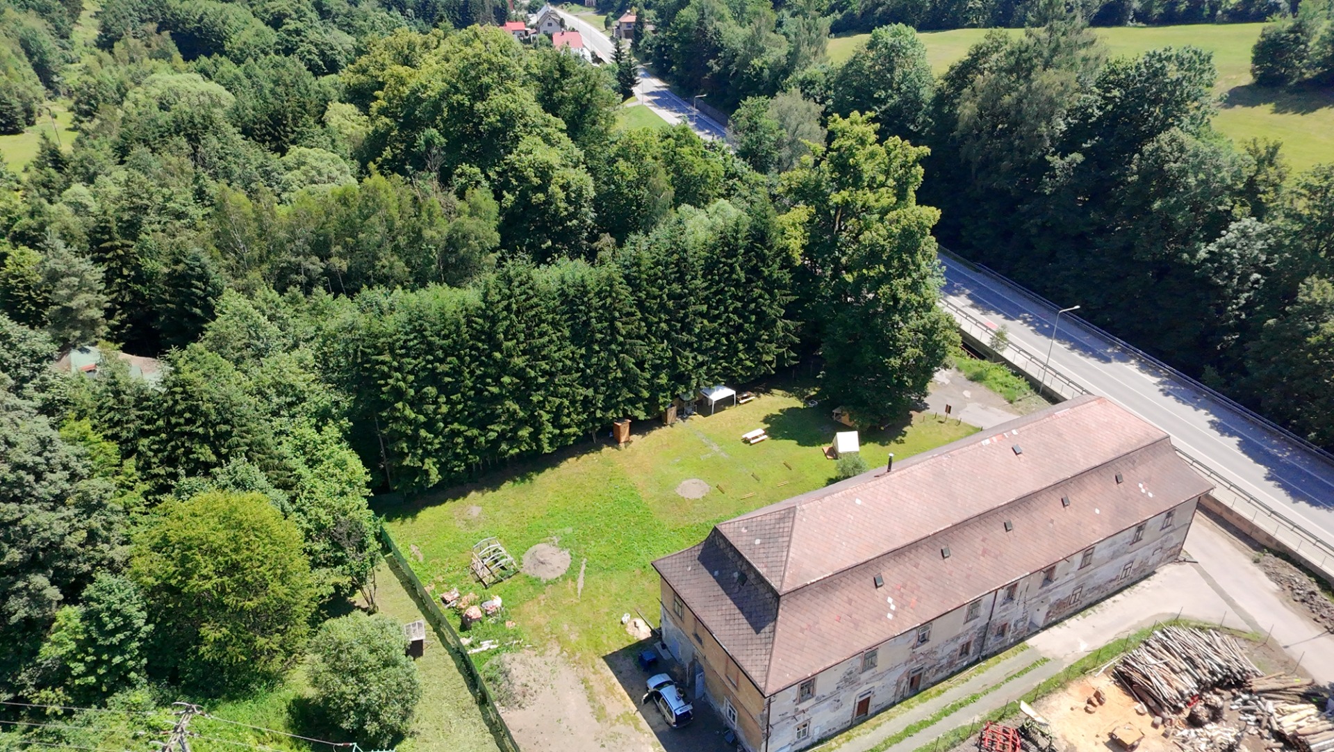 Letecký pohled na silnici uprostřed zeleného lesa, budovy a dvůr s trávníkem.